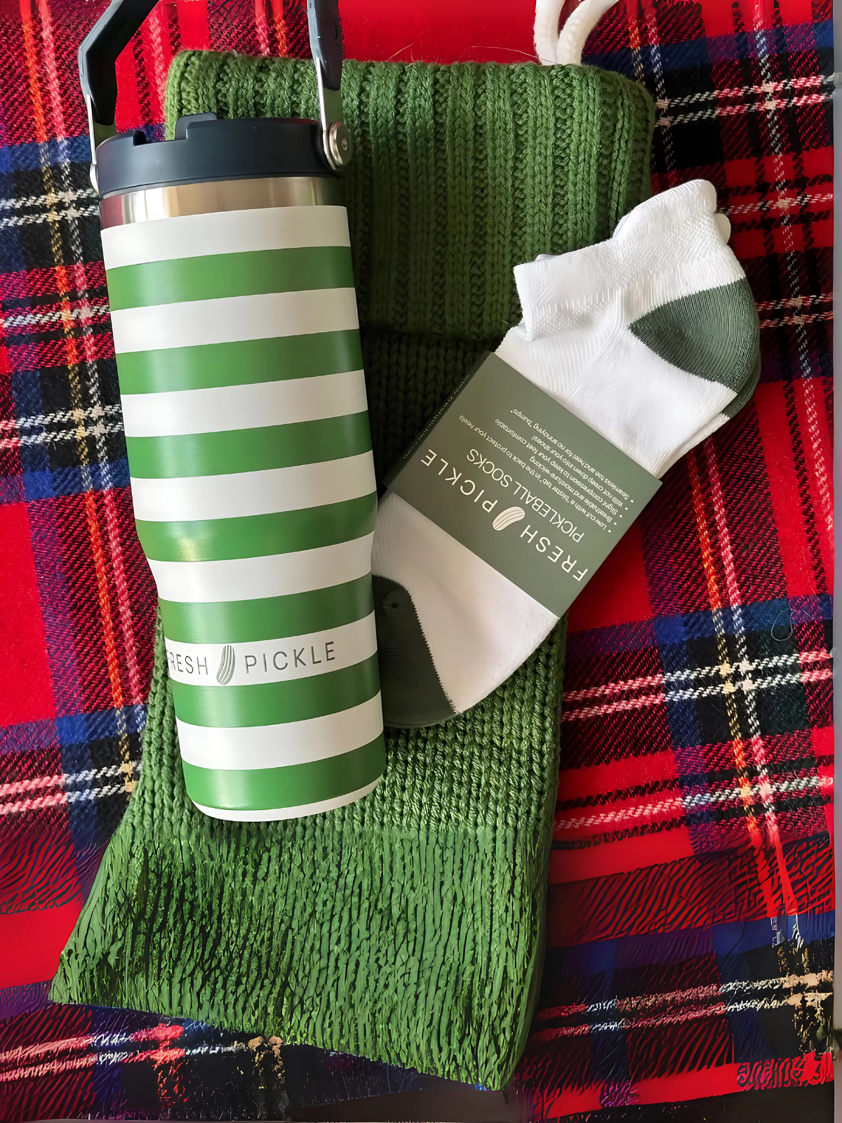 Green striped pickleball tumbler, green knit scarf, and socks on a red plaid blanket