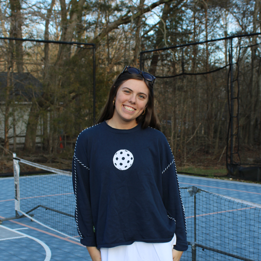Woman in navy pickleball sweatshirt and white skirt standing on outdoor pickleball court