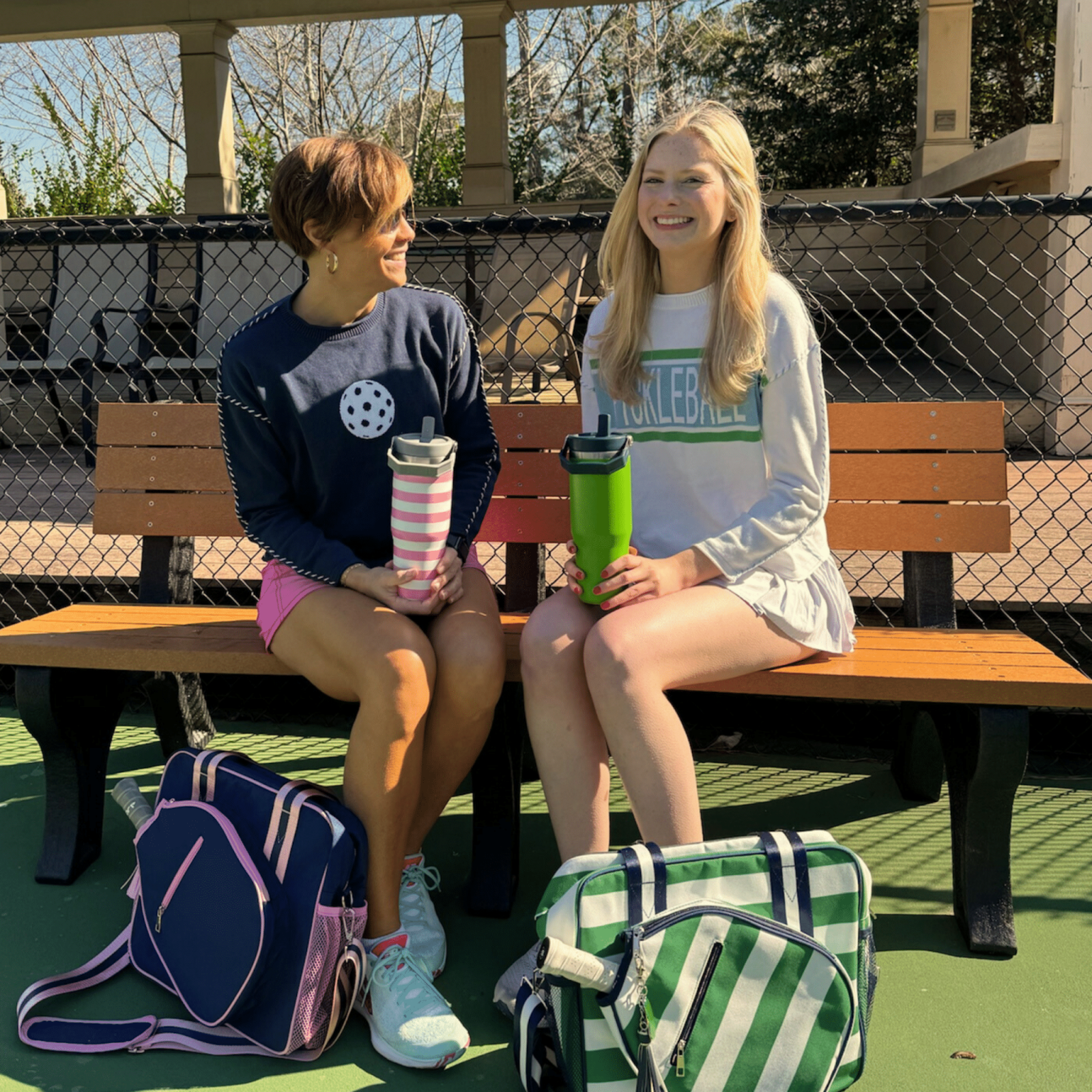 Two women with pickleball bags and water bottles sitting on a bench at an outdoor pickleball court
