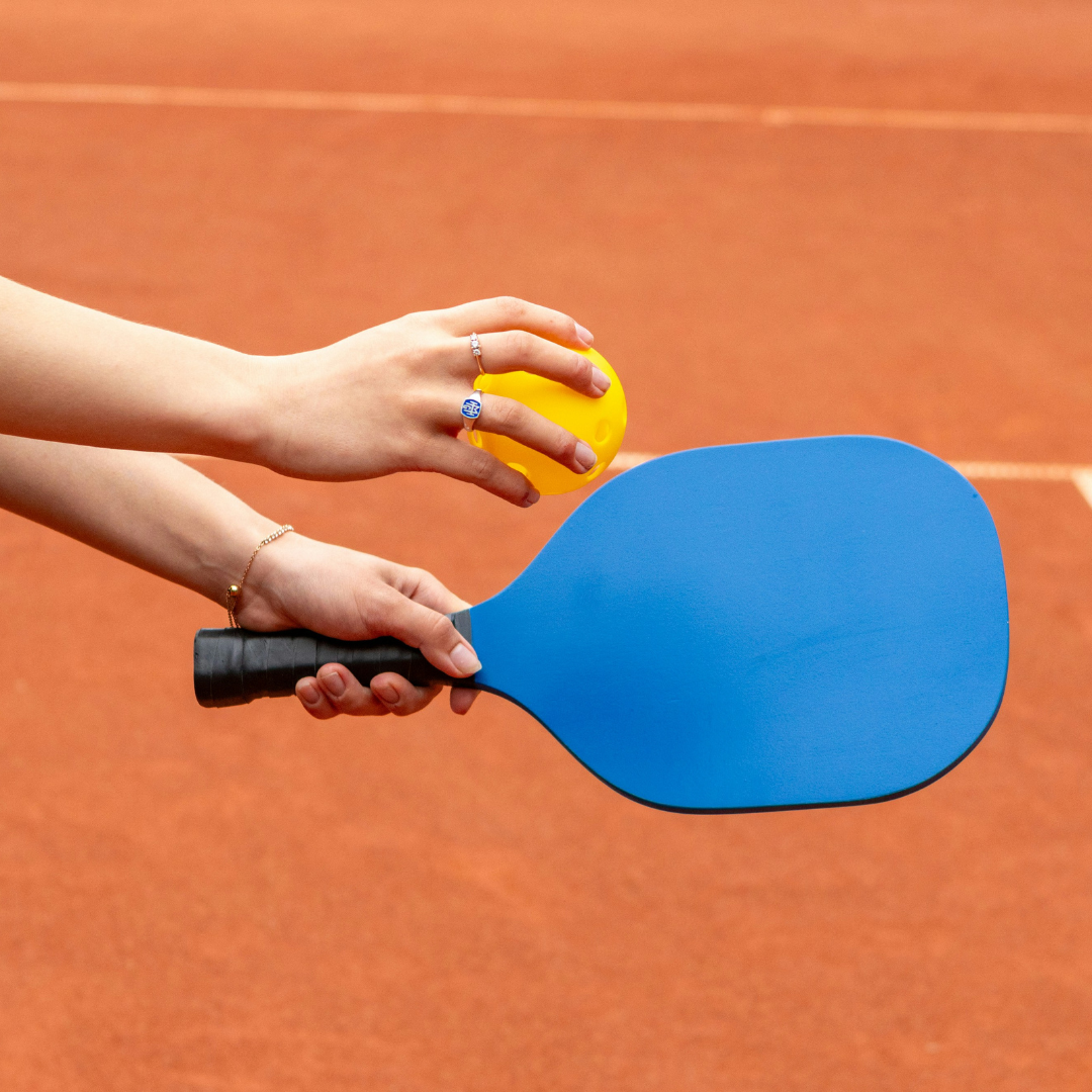 Hand holding blue pickleball paddle and yellow pickleball on outdoor court