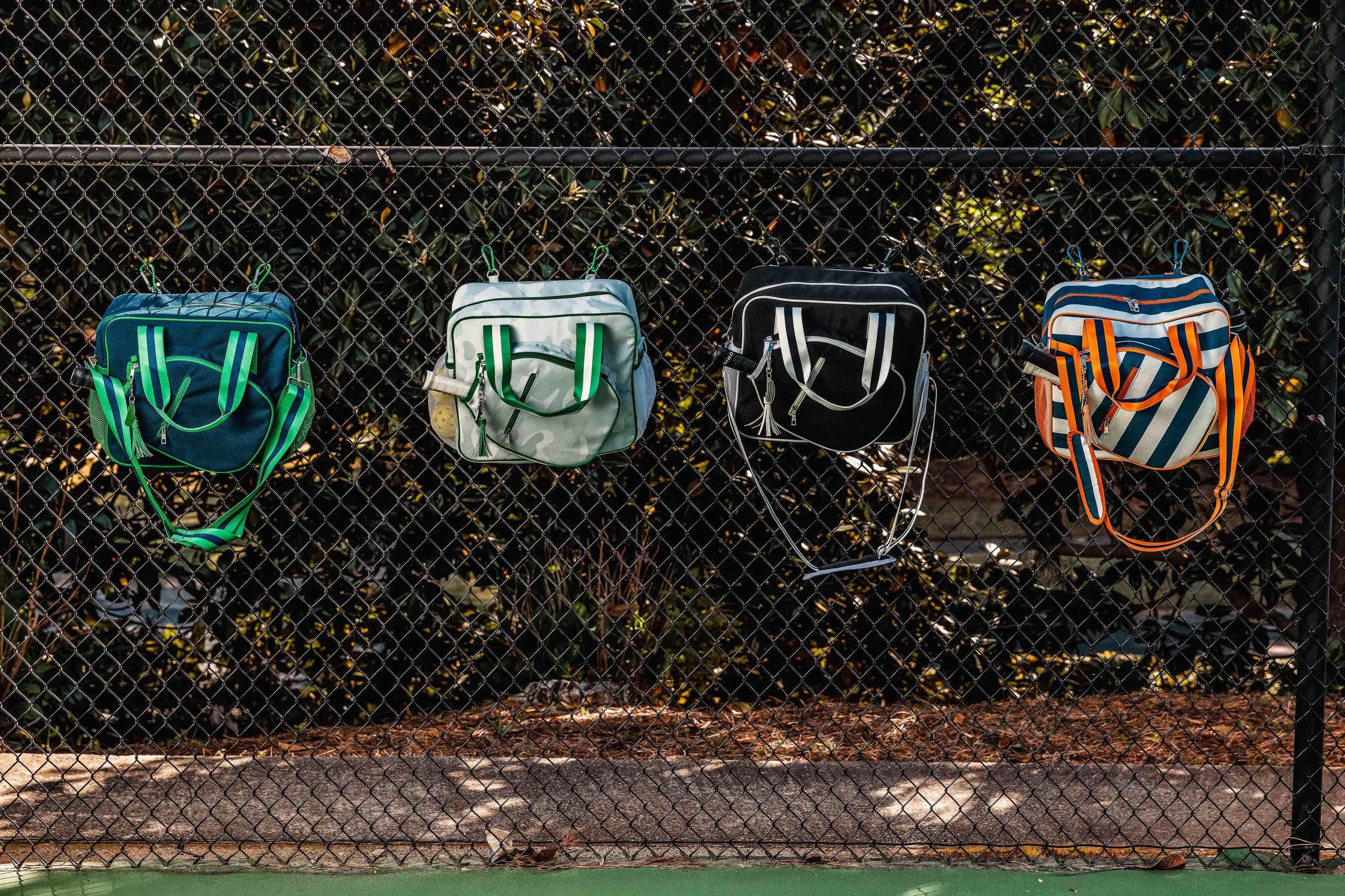 Four colorful pickleball bags hanging on a chain-link fence at an outdoor court