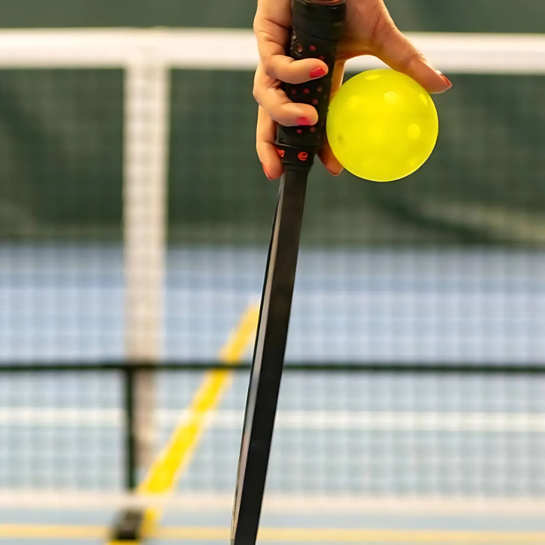 Hand holding a pickleball paddle and yellow pickleball on an indoor court
