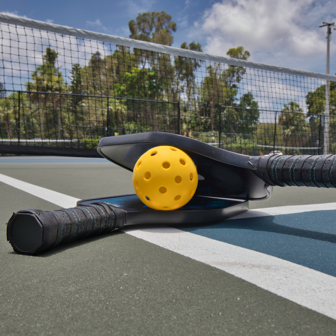 Pickleball paddles and yellow ball on outdoor court with net in background