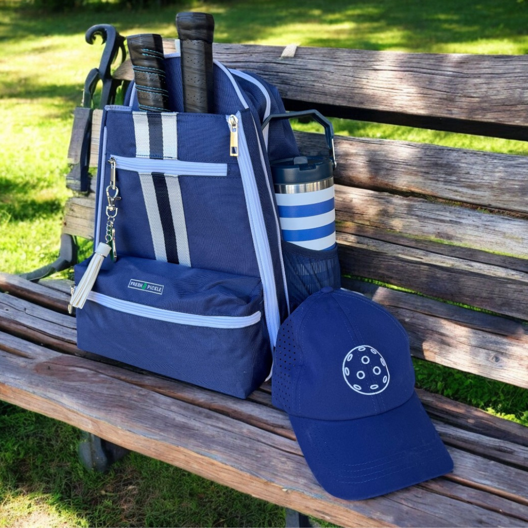 Blue pickleball bag with paddles, water bottle, and pickleball cap on a park bench