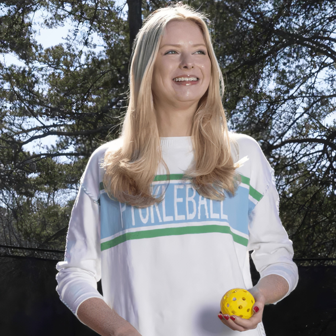 Woman outdoors in a pickleball sweatshirt holding a yellow pickleball, trees in background