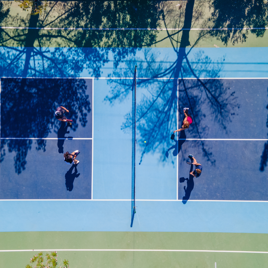 Aerial view of four people playing doubles pickleball on an outdoor court with tree shadows.