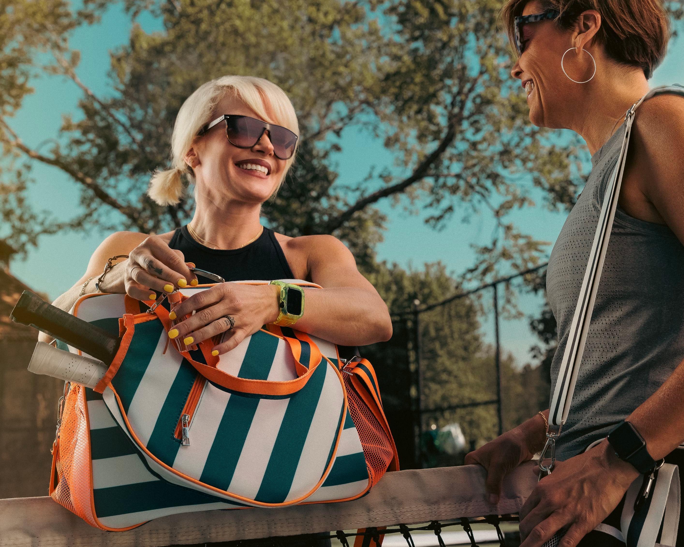 Two women with pickleball bags and paddles talking by a court on a sunny day