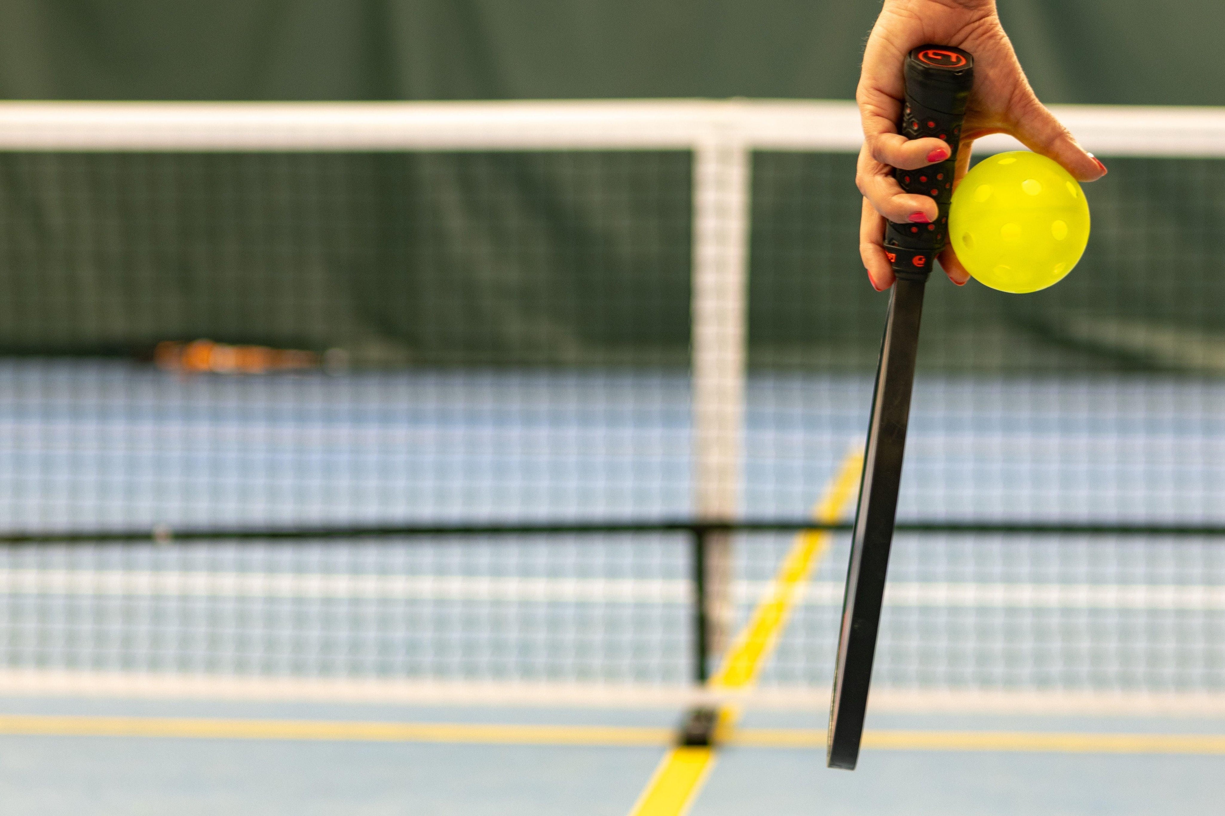 Hand holding a pickleball paddle and yellow pickleball on an indoor court