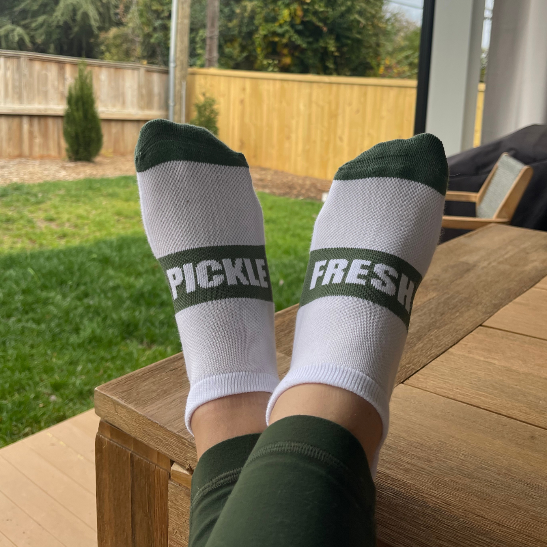 White and green pickleball socks with 'FRESH' and 'PICKLE' text, worn outdoors on a patio.