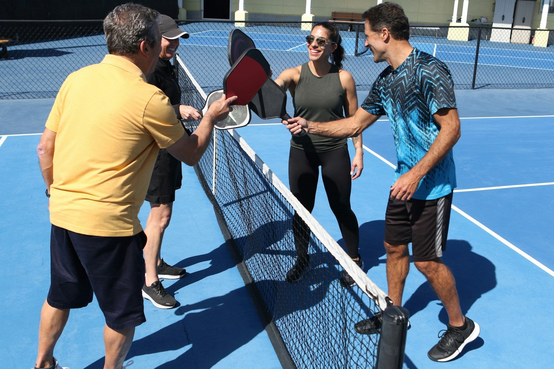 Four adults on outdoor pickleball court tapping paddles over the net, smiling