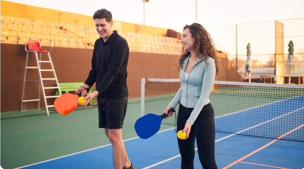 Two people playing pickleball on an outdoor court, holding paddles and yellow pickleballs.