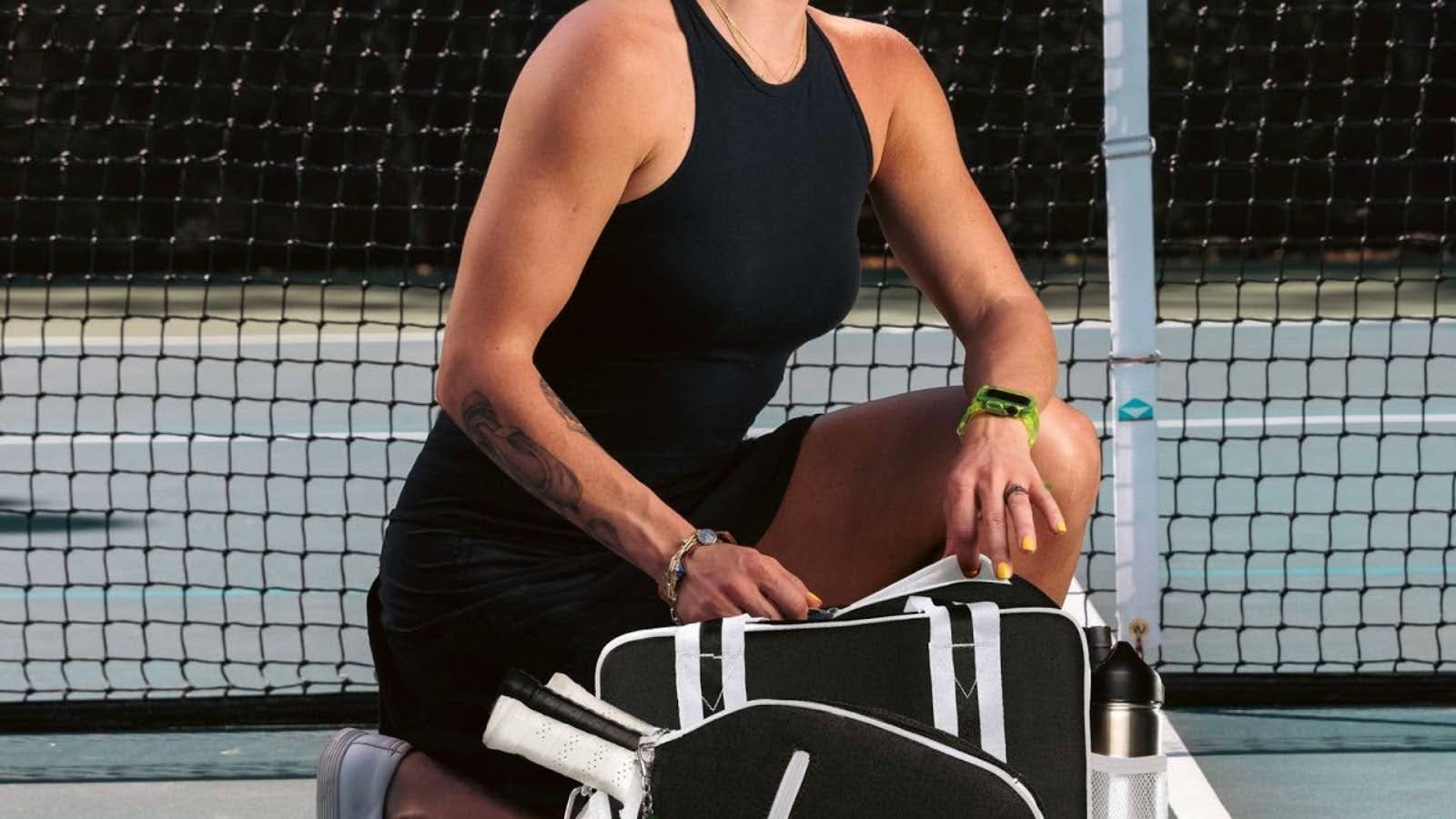 Woman in athletic dress kneeling on pickleball court with black pickleball bag and paddles
