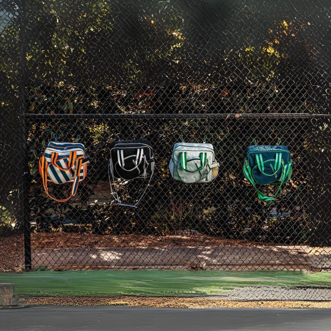Four colorful pickleball bags hanging on a chain link fence at an outdoor court.