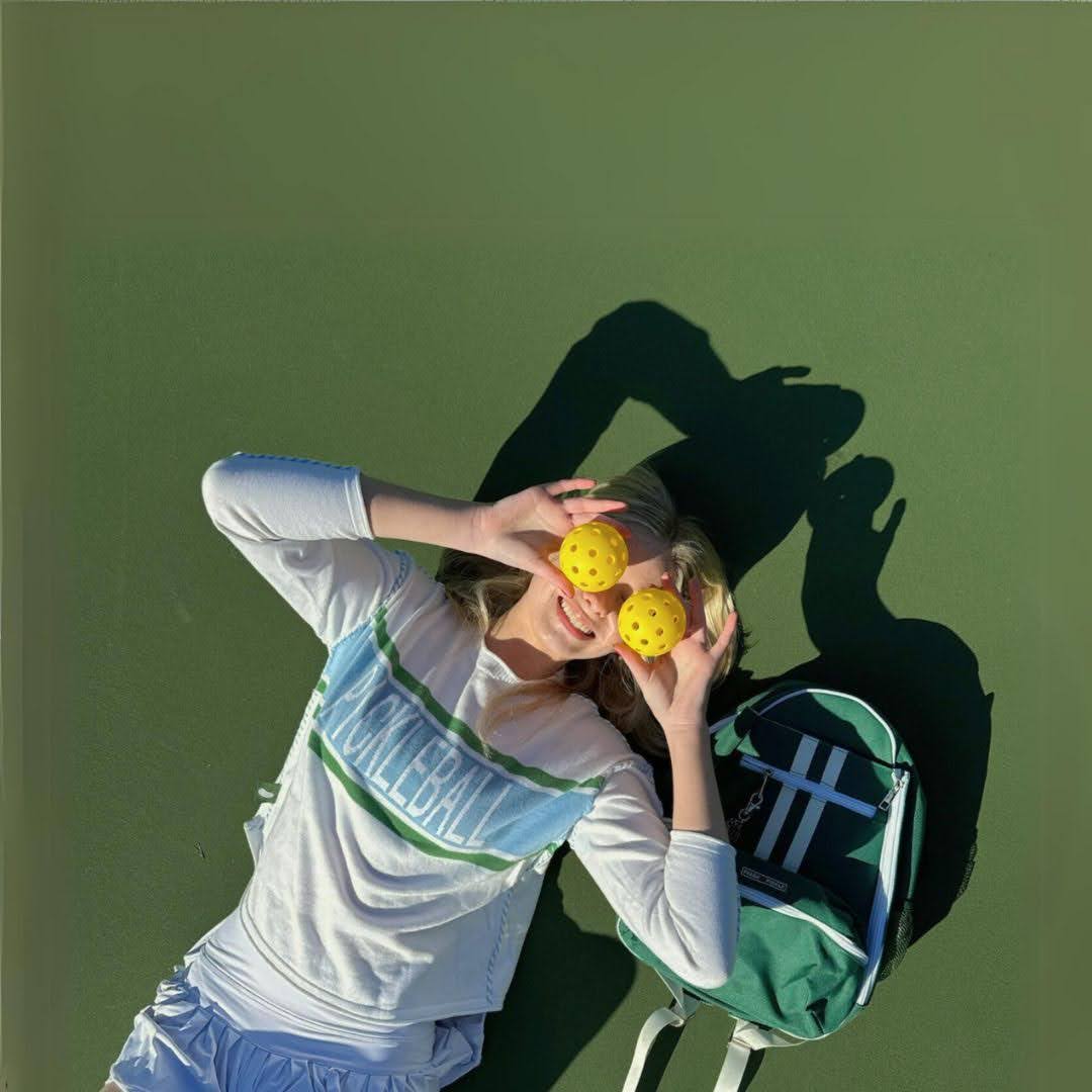 Woman on pickleball court holding yellow balls over eyes, wearing pickleball shirt, green bag nearby
