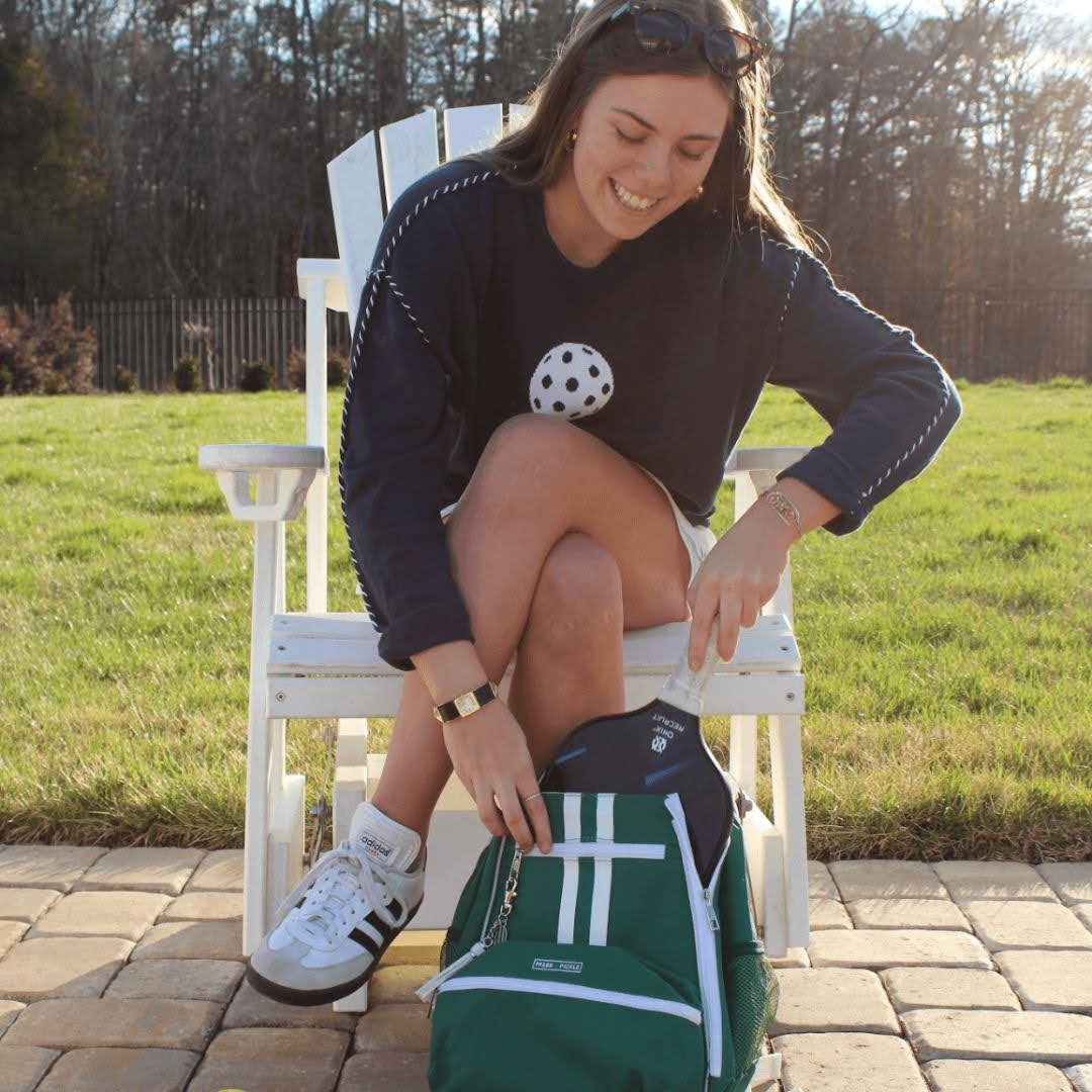 Woman sitting outdoors in athletic wear placing pickleball paddle in green pickleball bag