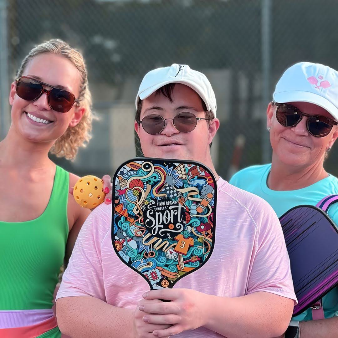 Three people with sunglasses holding colorful pickleball paddles and a pickleball on a court