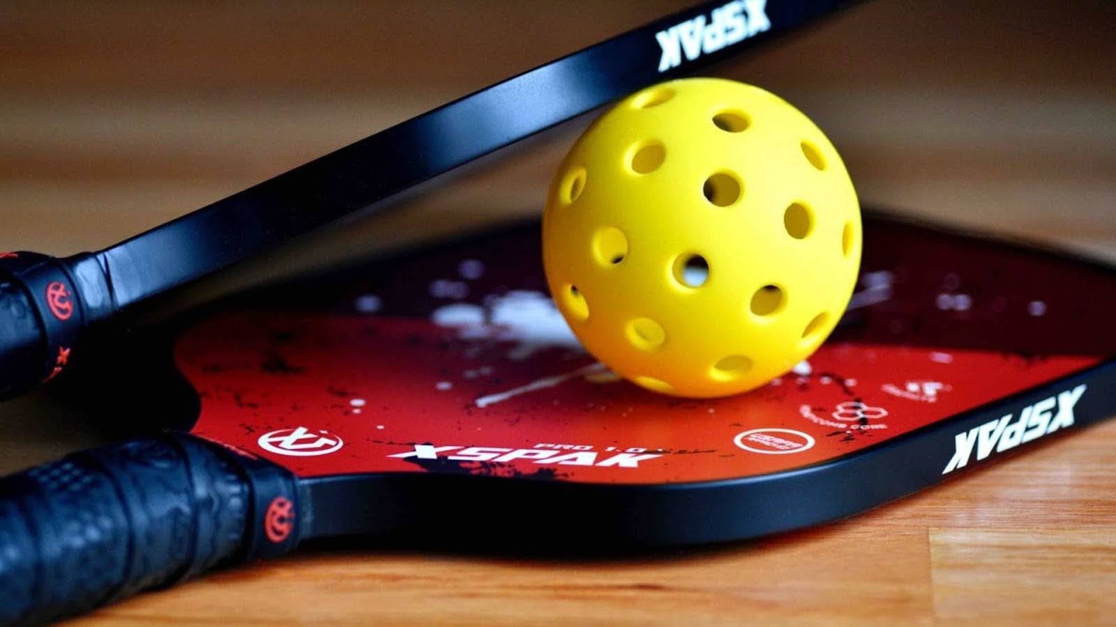 Yellow pickleball and two red pickleball paddles on wood floor, close-up view