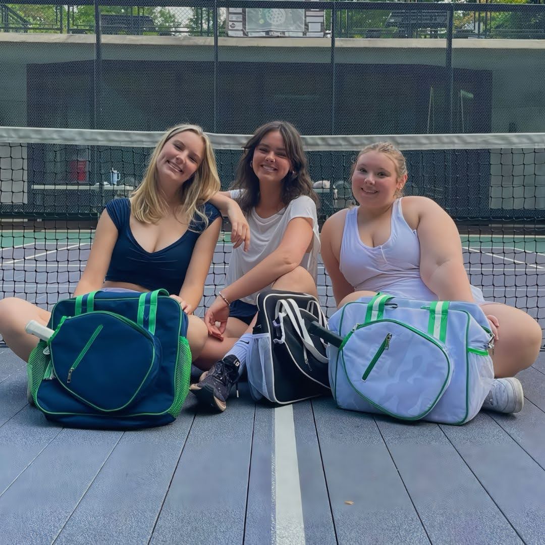 Three women sitting on a pickleball court with stylish pickleball gear bags and paddles