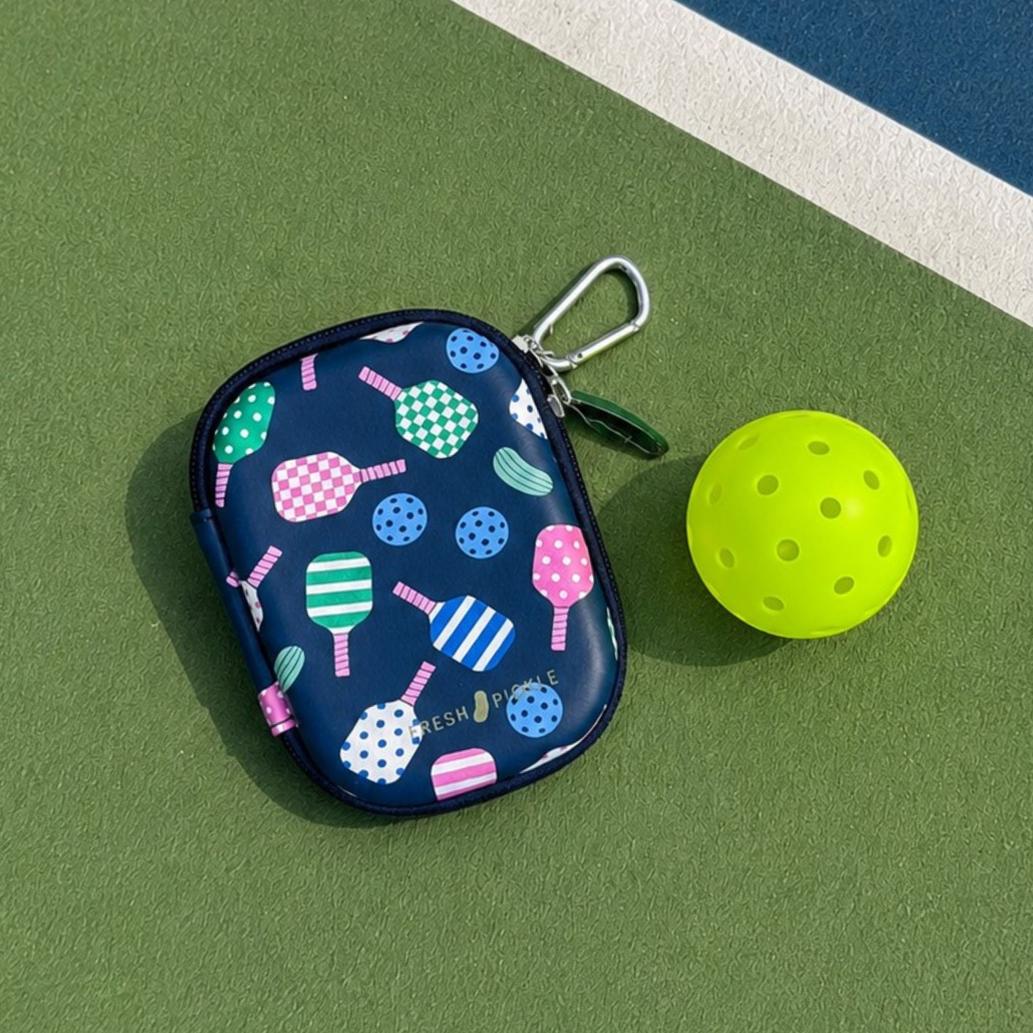 A navy blue zippered pouch with a colorful pickleball-themed print of rackets, balls, and a carabiner, placed on a green pickleball court next to a yellow pickleball.