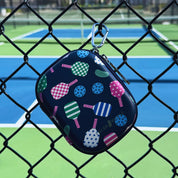 A small zippered case with a colorful pickleball pattern, hanging from a silver carabiner on a chain-link fence in front of a pickleball court.