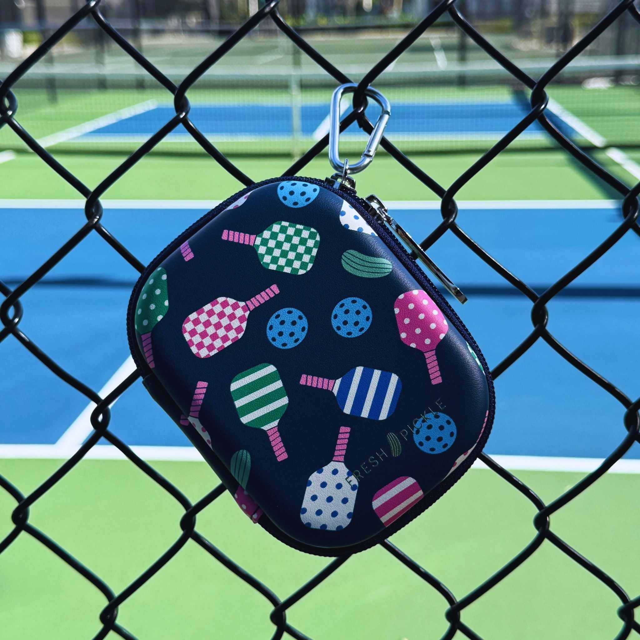 A small zippered case with a colorful pickleball pattern, hanging from a silver carabiner on a chain-link fence in front of a pickleball court.